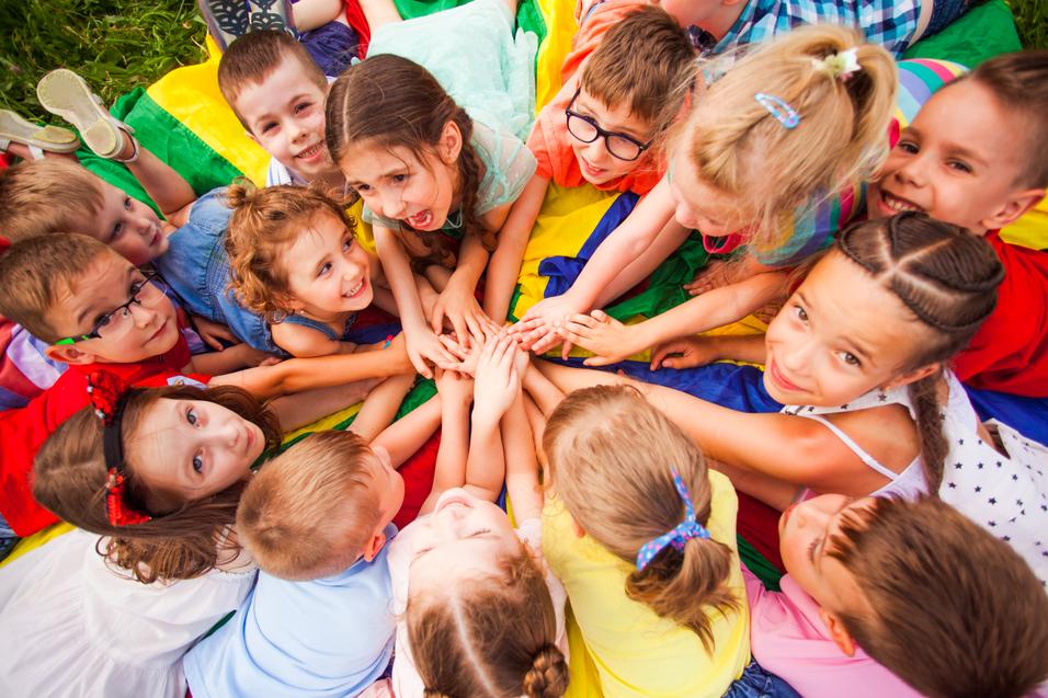 Eine Gruppe von Kindern sitzt im Kreis und legt ihre H&auml;nde aufeinander, umgeben von buntem Tuch.