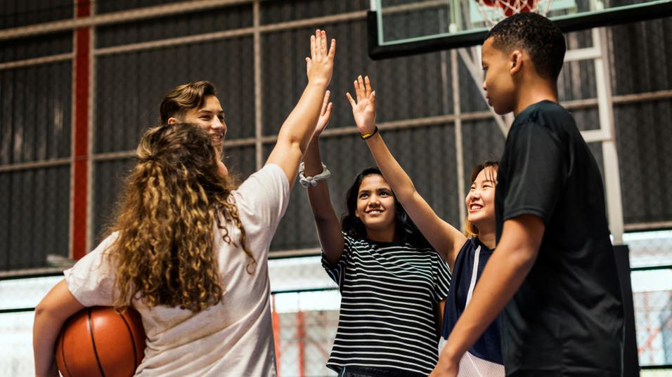 Vier Jugendliche feiern mit erhobenen H&auml;nden nach einem Basketballspiel in einer Sporthalle. Ein Ball liegt in der N&auml;he.