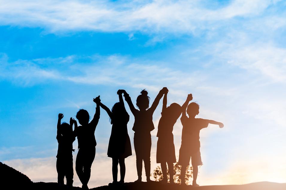 Silhouette von sechs Kindern, die bei Sonnenuntergang jubelnd die Arme heben, vor einem blauen Himmel.