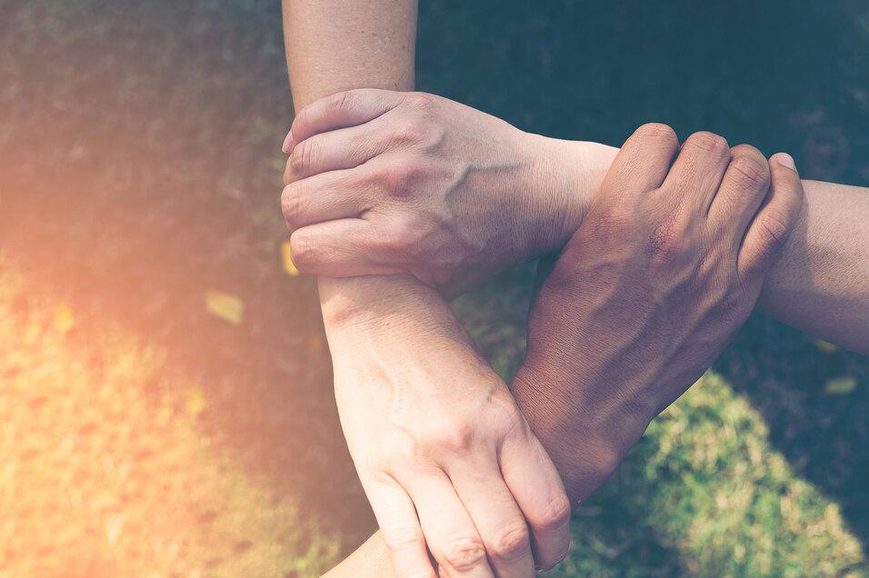 Vier H&auml;nde greifen ineinander auf einer gr&uuml;nen Wiese, symbolisieren Zusammenhalt und Kooperation.