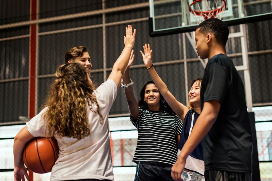 Vier Jugendliche feiern mit erhobenen H&auml;nden nach einem Basketballspiel in einer Sporthalle. Ein Ball liegt in der N&auml;he.