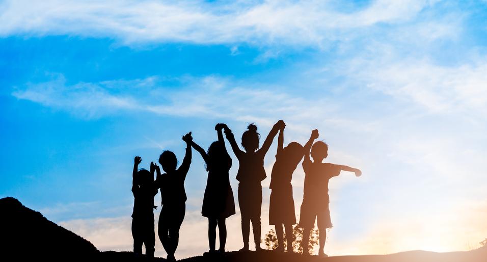 Silhouette von sechs Kindern, die bei Sonnenuntergang jubelnd die Arme heben, vor einem blauen Himmel.