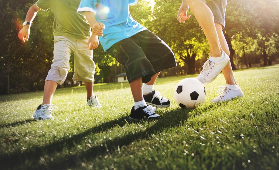 Drei Kinder spielen auf einer Wiese Fu&szlig;ball, w&auml;hrend ein Ball im Vordergrund zwischen ihnen liegt.