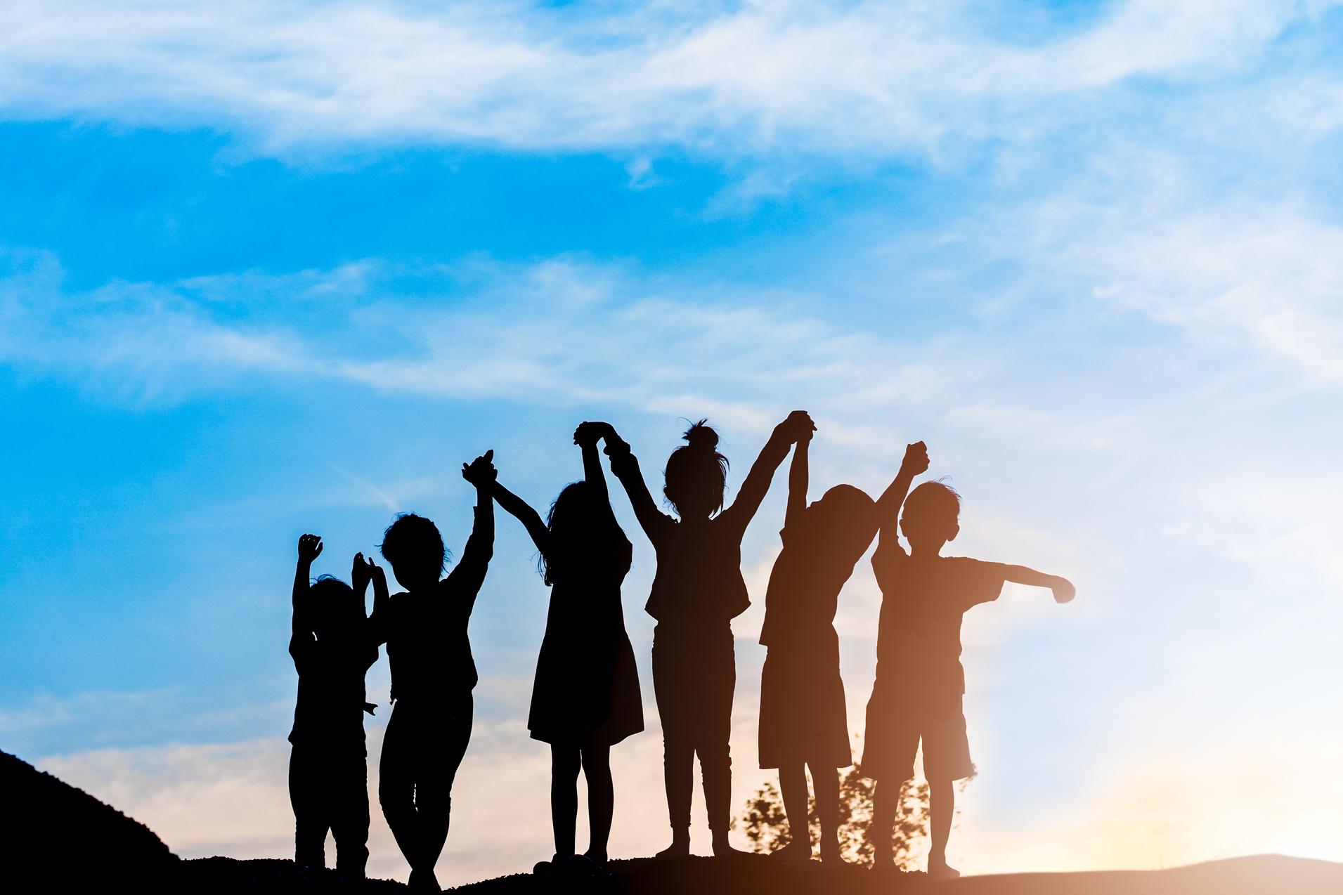 Silhouette von sechs Kindern, die bei Sonnenuntergang jubelnd die Arme heben, vor einem blauen Himmel.