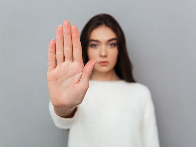 Junge Frau in wei&szlig;em Pullover h&auml;lt ihre Hand mit ausgestreckten Fingern in die Kamera. Hintergrund in Grau.