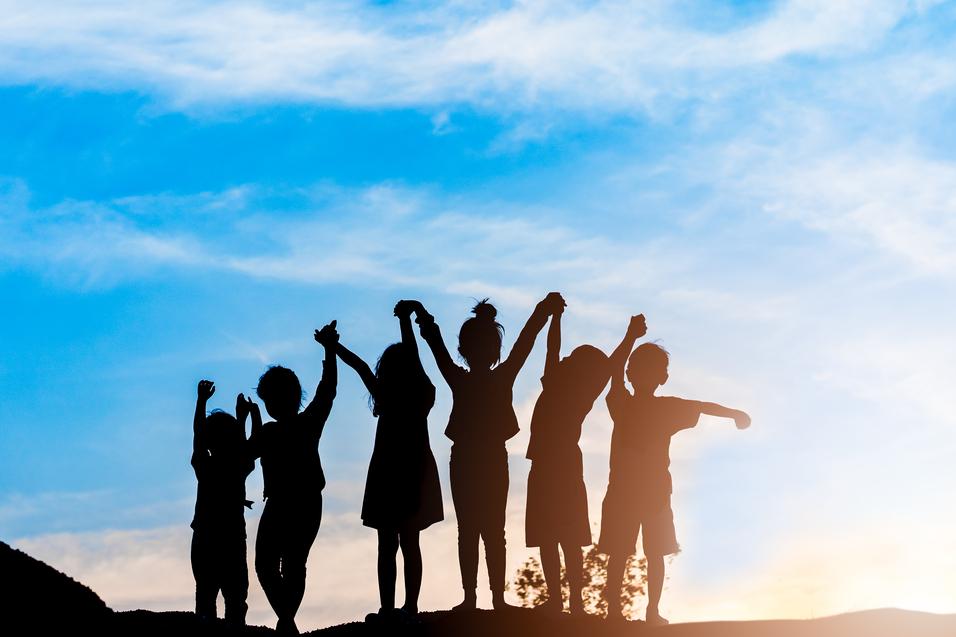 Silhouette von sechs Kindern, die bei Sonnenuntergang jubelnd die Arme heben, vor einem blauen Himmel.