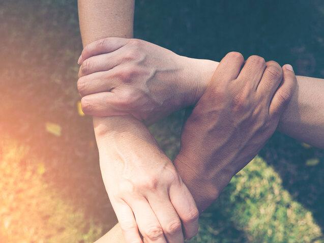 Vier H&auml;nde greifen ineinander auf einer gr&uuml;nen Wiese, symbolisieren Zusammenhalt und Kooperation.