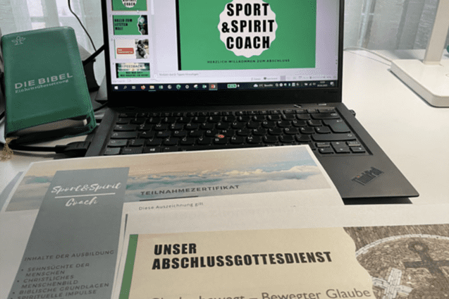 Laptop mit Pr&auml;sentation "Sport & Spirit Coach" und Teilnahmezertifikat, daneben Die Bibel und Unterlagen auf einem Tisch.