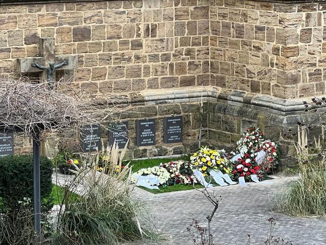 Blumenarrangements und Gedenktafeln stehen an einer Steinmauer in einer ruhigen Gartenfl&auml;che einer Kirche.