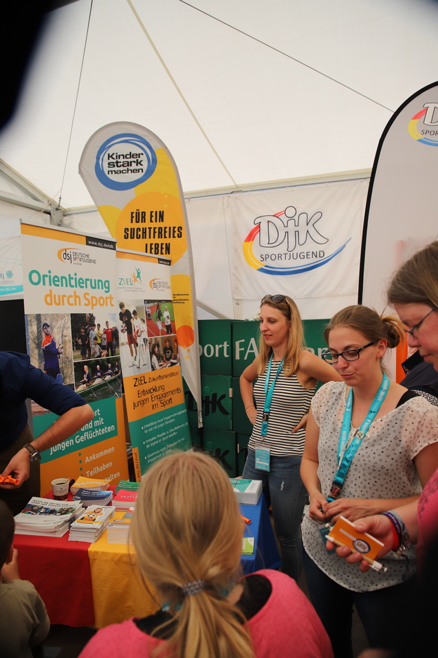 Zelt mit Informationsst&auml;nden und jungen Erwachsenen, die &uuml;ber Sport und Integration diskutieren. Plakate informieren &uuml;ber Programme.