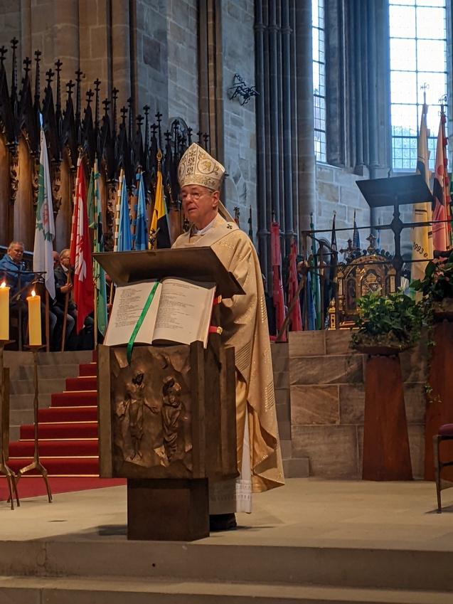 Mann in liturgischer Kleidung und Mitra steht an einer Pultb&uuml;hne in einer Kirche und h&auml;lt eine Ansprache.