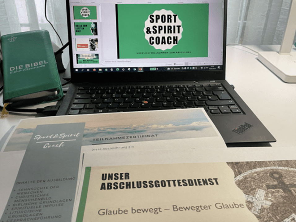 Laptop mit Pr&auml;sentation "Sport & Spirit Coach" und Teilnahmezertifikat, daneben Die Bibel und Unterlagen auf einem Tisch.