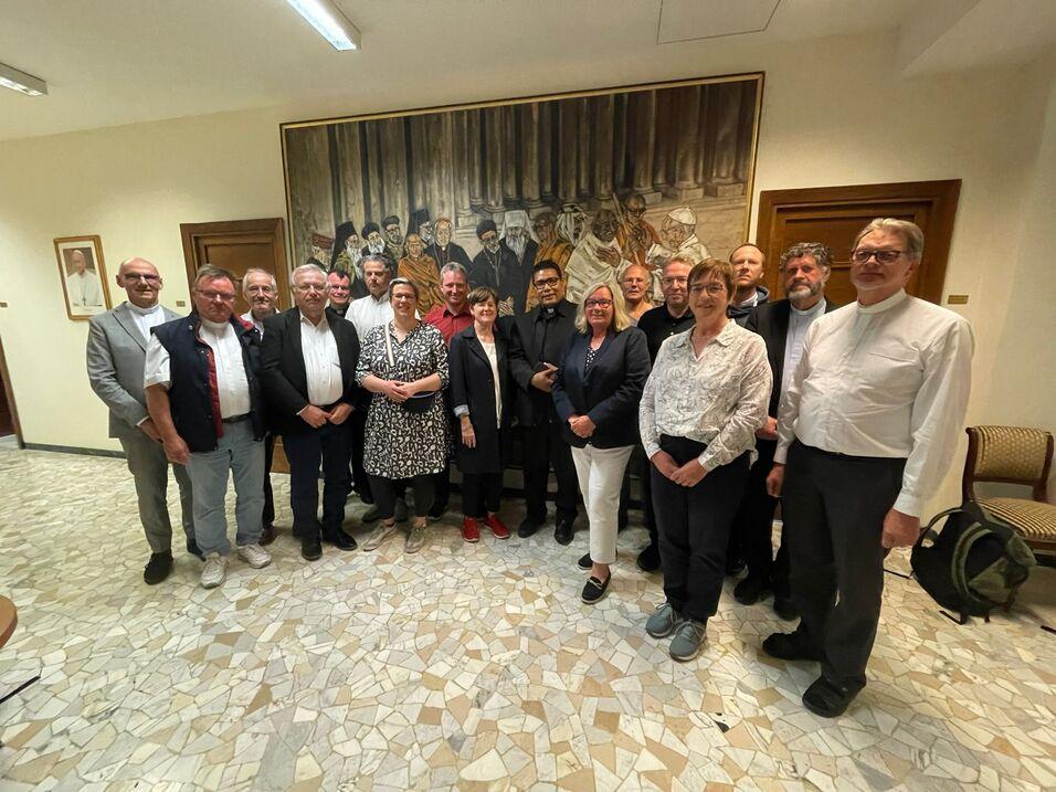 Gruppennahaufnahme von 16 Personen vor einem Wandgem&auml;lde in einem Innenraum mit Fliesenboden.