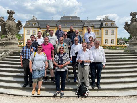 Gruppe von 20 Personen steht auf einer Treppe vor einem historischen Geb&auml;ude mit Garten und Wolkenhimmel.