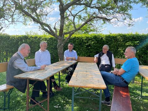 F&uuml;nf M&auml;nner sitzen um eine rustikale Holztischgruppe im Freien, umgeben von B&auml;umen und gr&uuml;nem Gestr&uuml;pp.