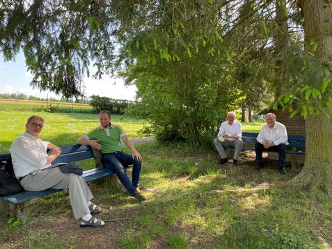 Vier M&auml;nner sitzen auf einer Bank im Schatten von B&auml;umen, umgeben von einer Wiese und einem l&auml;ndlichen Hintergrund.
