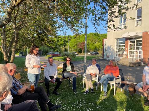Gruppenszenario im Freien mit mehreren Personen, die an einem Gespr&auml;ch teilnehmen, w&auml;hrend sie auf St&uuml;hlen unter einem Baum sitzen.