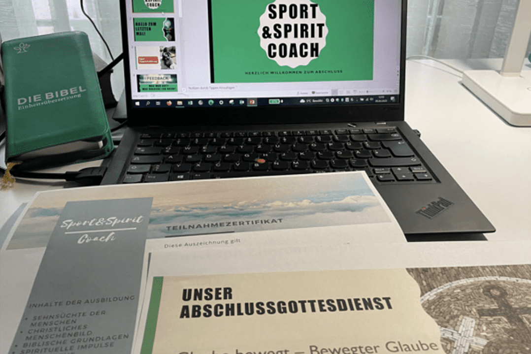 Laptop mit Pr&auml;sentation "Sport & Spirit Coach" und Teilnahmezertifikat, daneben Die Bibel und Unterlagen auf einem Tisch.