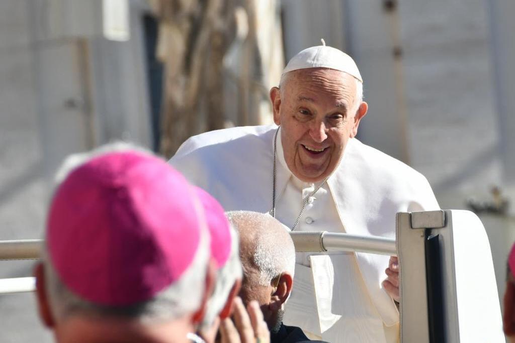 Papst Franziskus l&auml;chelt und winkt von einem Podium aus, w&auml;hrend Bisch&ouml;fe im Hintergrund zu sehen sind.