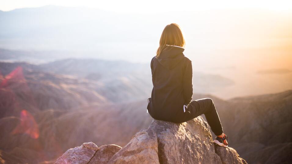 Frau sitzt auf einem Felsen und blickt auf eine weite, bergige Landschaft bei Sonnenaufgang.