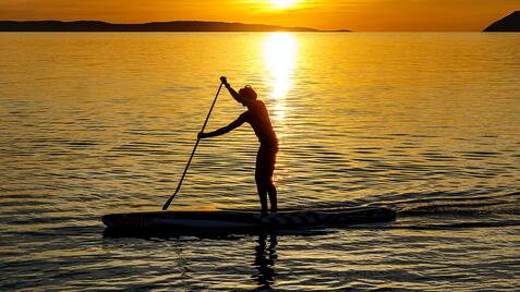 Person steht auf einem Stand-Up-Paddle-Board und paddelt bei Sonnenuntergang auf ruhigem Wasser.