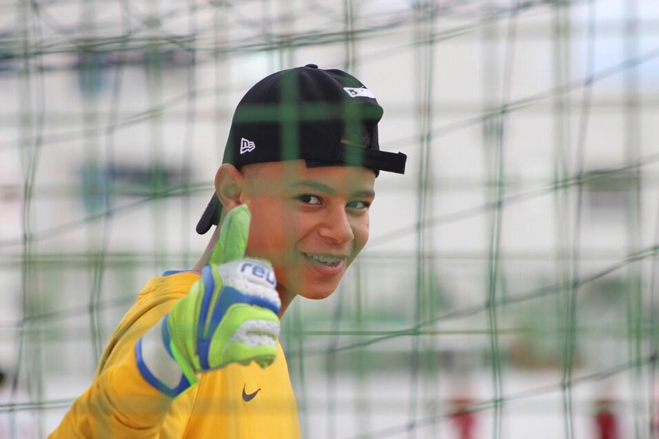 Junger Torwart mit Handschuhen und Cap gibt Daumen hoch hinter einem Tornetz.