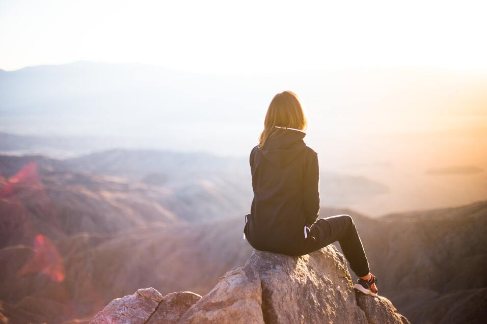 Frau sitzt auf einem Felsen und blickt auf eine weite, bergige Landschaft bei Sonnenaufgang.