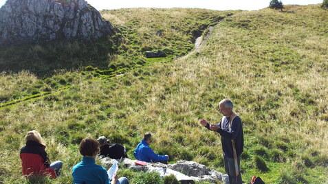Eine Gruppe von Menschen sitzt auf einer Wiese, w&auml;hrend ein &auml;lterer Mann im Vordergrund spricht.