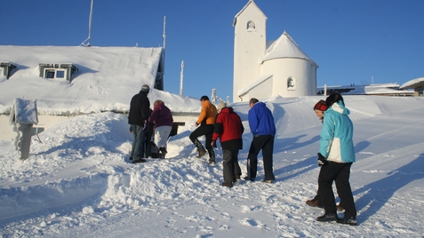 Gruppe von Personen, die durch hohen Schnee in Richtung einer Kirche mit wei&szlig;em Turm gehen, blauer Himmel im Hintergrund.