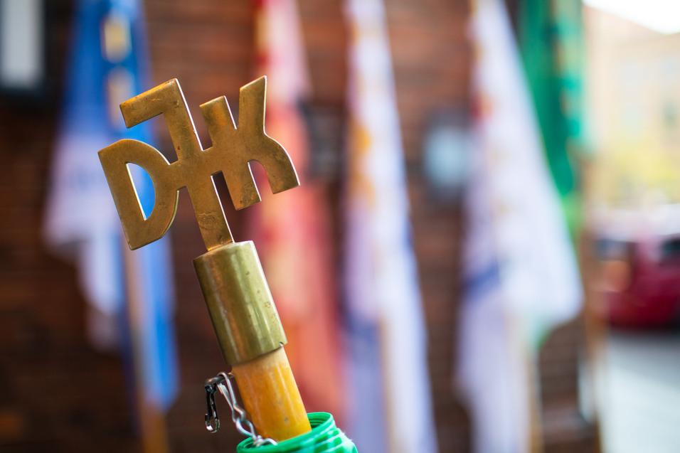 Nahaufnahme eines goldenen Schl&uuml;ssels mit dem Schriftzug &bdquo;DJK&ldquo; vor unscharfen Fahnen im Hintergrund.