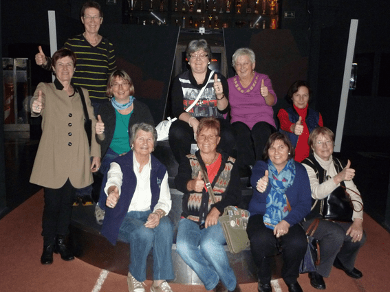 Gruppe von zehn Frauen, die auf einer Treppe sitzen und alle Daumen hoch zeigen, hinter ihnen eine dunkle Wand.