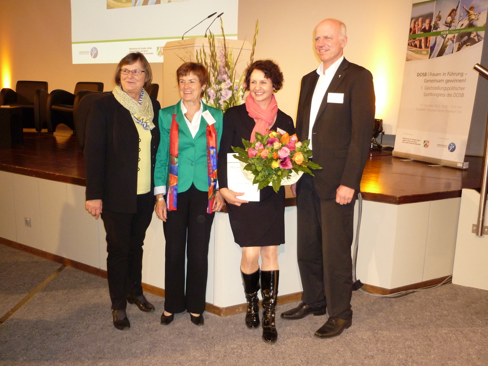 Vier Personen stehen auf einer B&uuml;hnenfl&auml;che, eine h&auml;lt einen Blumenstrau&szlig;, w&auml;hrend die anderen l&auml;cheln und applaudieren.
